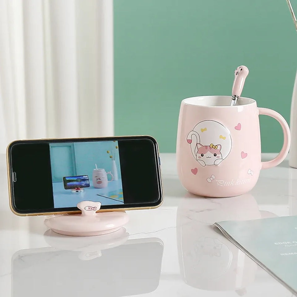 une jolie tasse rose au design d'un adorable chat. La tasse possède un couvercle doté d'un poisson et d'une cuillère en forme de patte de chat. Le couvercle sert également de support pour smartphone. Vous pouvez y mettre votre smartphone pour regarder une série pendant que vous savourez un café.