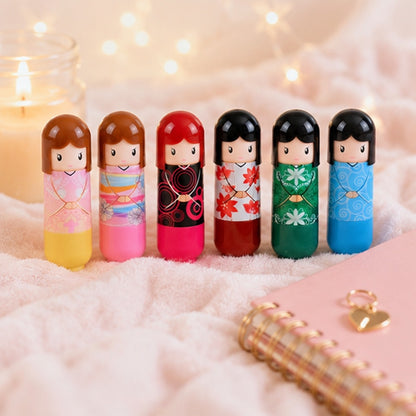 Ce sont des baumes à lèvres en forme de petits personnage kokeshi. dans le style kawaii japonais. ils sont posés debout sur une couverture blanche douce.
