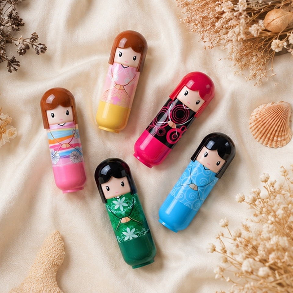 Ce sont des baumes à lèvres en forme de petits personnage kokeshi. dans le style kawaii japonais. ils sont posés allongés sur une couverture blanche douce.