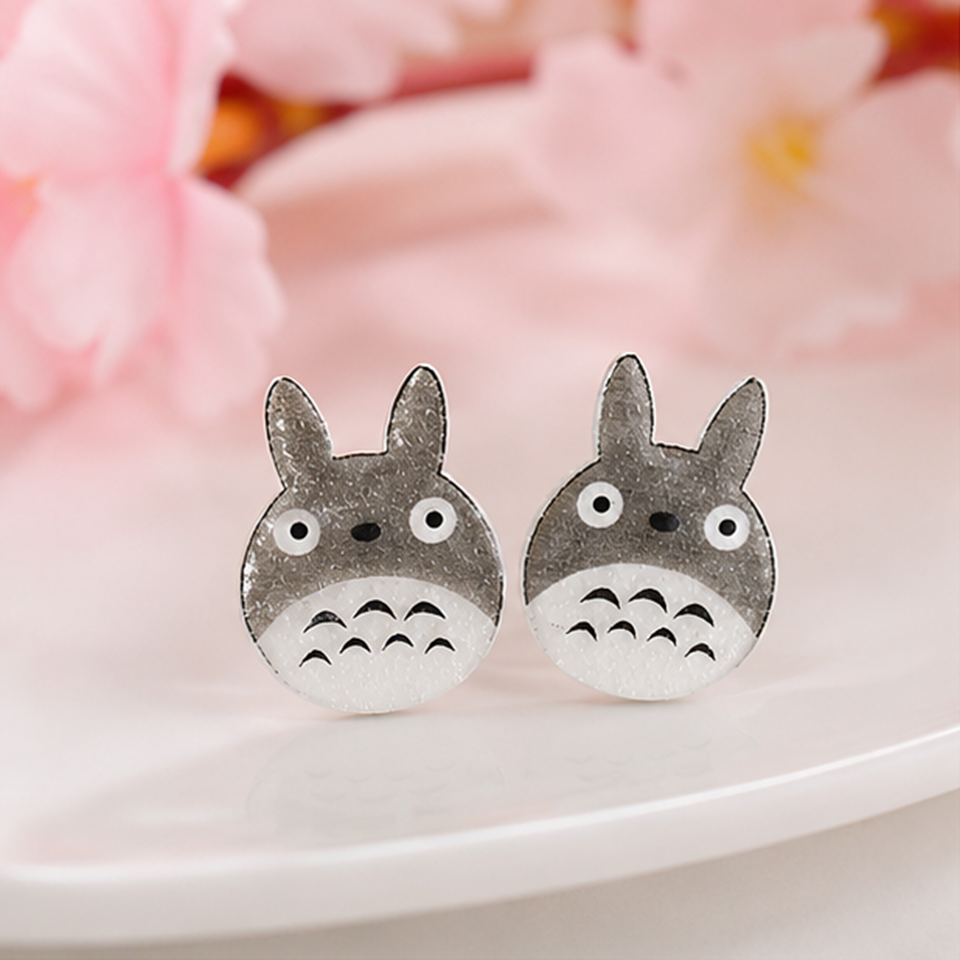 une paire de boucles d'oreilles en forme de Totoro façon chibi. le design est minimaliste kawaii mignon. les boucles d'oreilles sont pailletées.