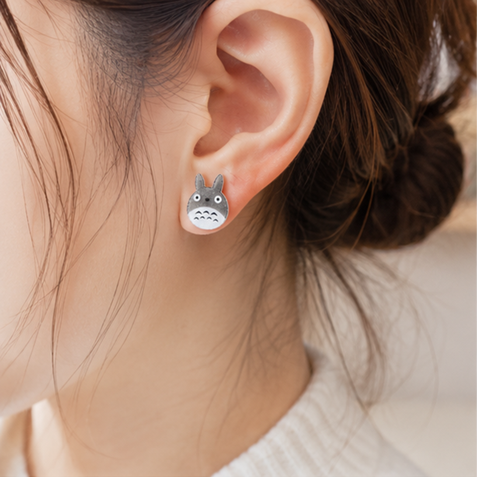 une paire de boucles d'oreilles en forme de Totoro façon chibi. le design est minimaliste kawaii mignon. les boucles d'oreilles sont pailletées.