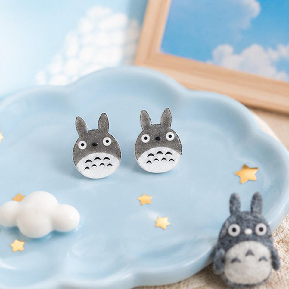 une paire de boucles d'oreilles en forme de Totoro façon chibi. le design est minimaliste kawaii mignon. les boucles d'oreilles sont pailletées.