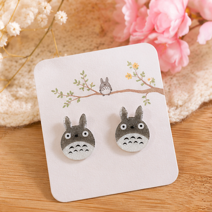 une paire de boucles d'oreilles en forme de Totoro façon chibi. le design est minimaliste kawaii mignon. les boucles d'oreilles sont pailletées.