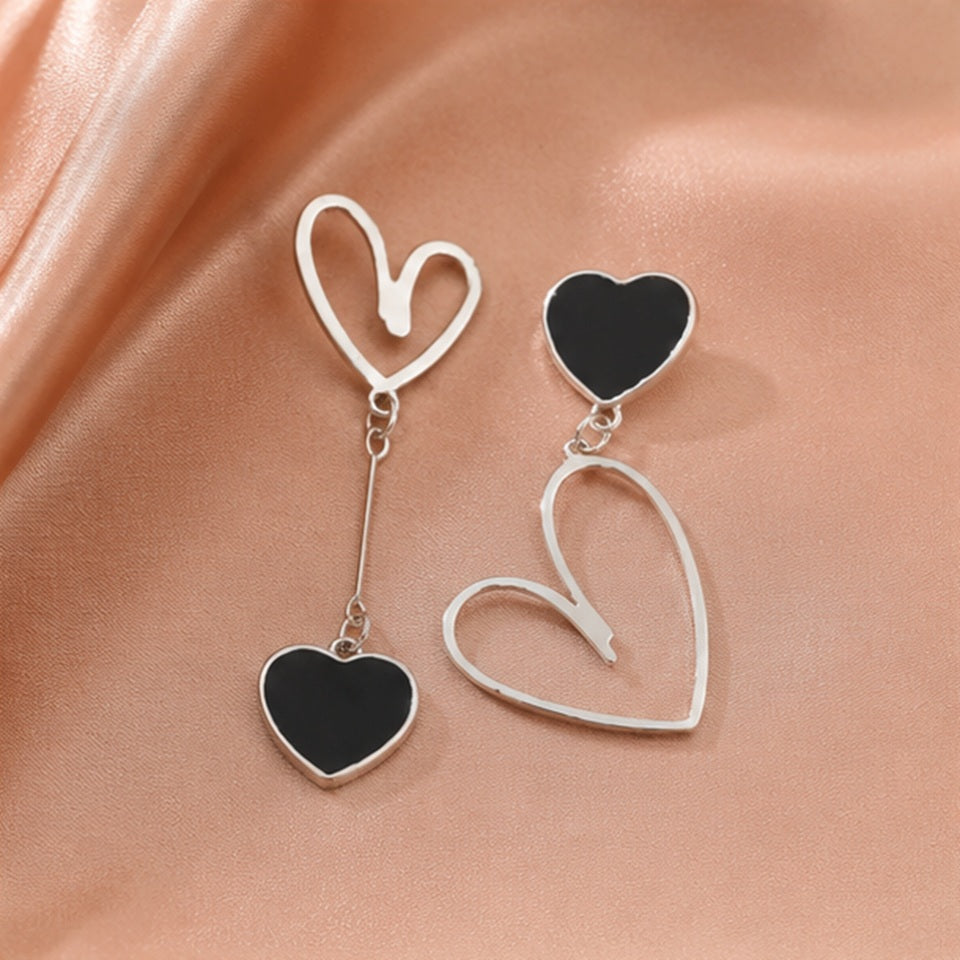 Boucles d’oreilles asymétriques cœurs noirs au style chic et moderne posées sur un tissu satiné rose ✨ Bijoux argentés avec cœurs noirs brillants et formes ouvertes minimalistes, parfaits pour ajouter une touche élégante, romantique et tendance à une tenue. Ambiance douce et raffinée inspirée de la mode kawaii adulte.