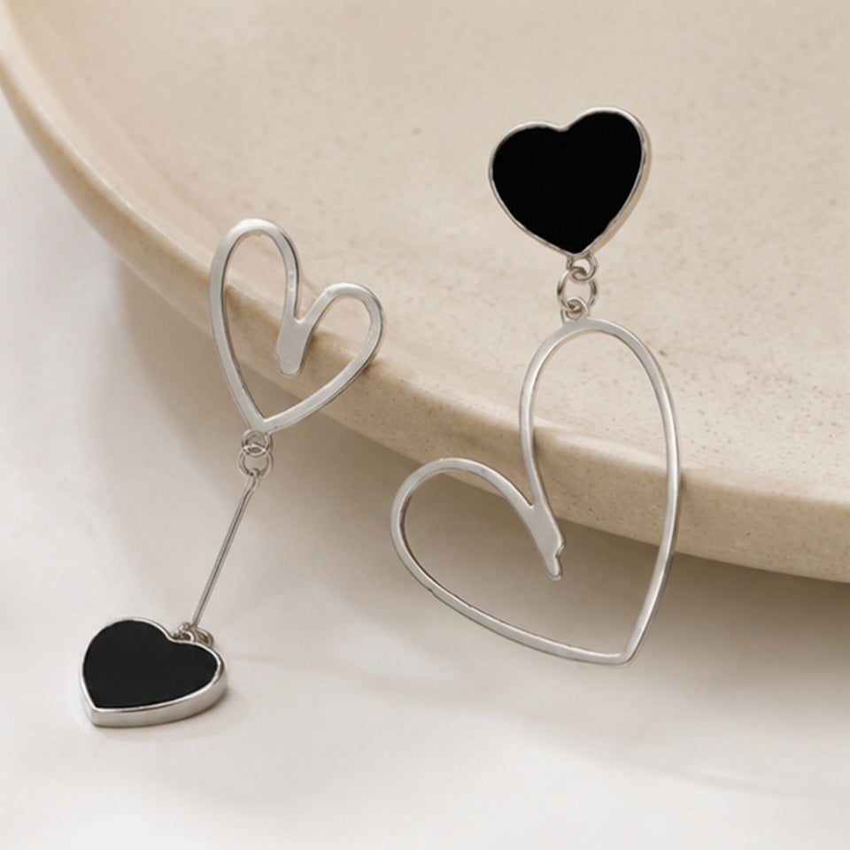 Boucles d’oreilles asymétriques cœurs noirs et argentés au design minimaliste et élégant ✨ Bijoux modernes avec cœurs ouverts argentés et pendentifs noirs brillants, parfaits pour ajouter une touche romantique, chic et tendance à une tenue. Ambiance douce et épurée avec décor beige raffiné.