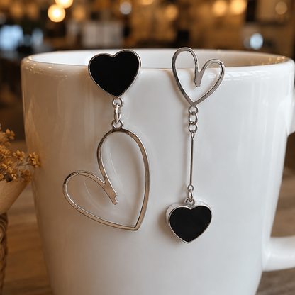 Boucles d’oreilles asymétriques cœurs noirs au design élégant et minimaliste ✨ Bijoux argentés avec cœurs noirs suspendus et formes ouvertes modernes, parfaits pour ajouter une touche chic, romantique et originale à une tenue. Ambiance chaleureuse et raffinée avec fond lumineux effet bokeh.