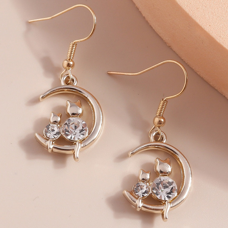 une paire de boucles d'oreilles en forme de lune dorée sur laquelle reposent deux adorables chats dont le corps est en strass brillant