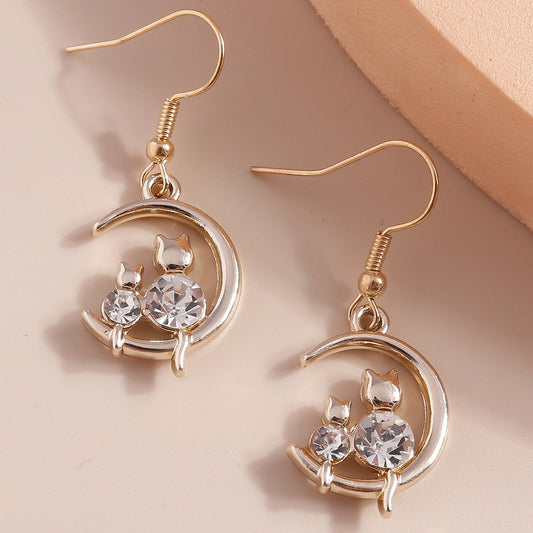 une paire de boucles d'oreilles en forme de lune dorée sur laquelle reposent deux adorables chats dont le corps est en strass brillant