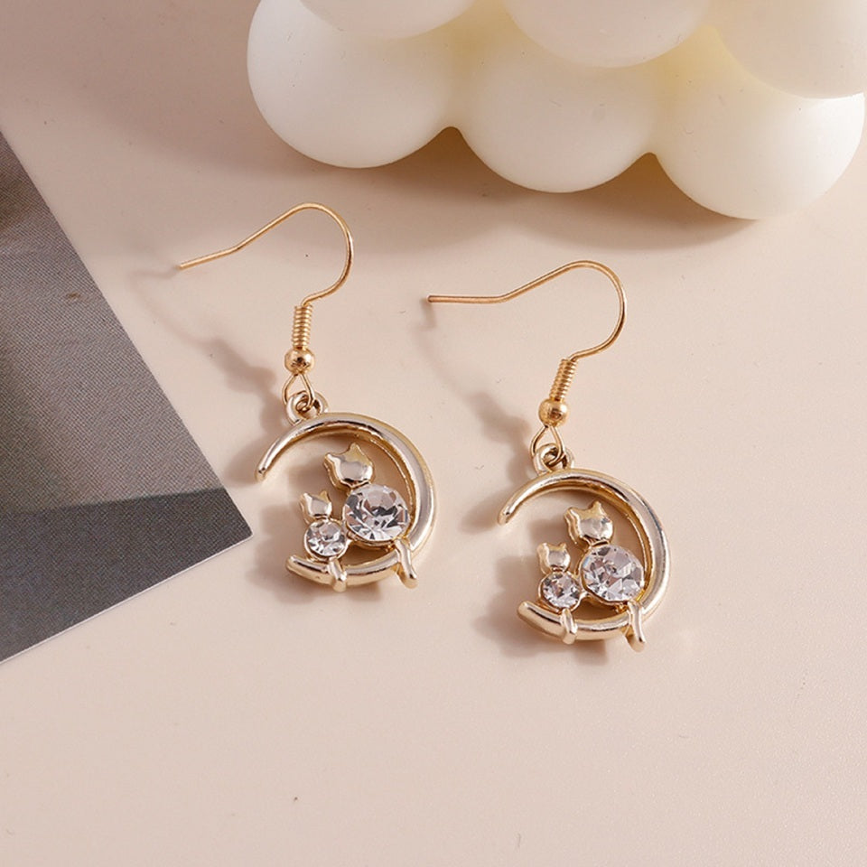une paire de boucles d'oreilles en forme de lune dorée sur laquelle reposent deux adorables chats dont le corps est en strass brillant