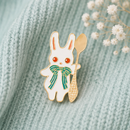 un pin's en forme de lapin blanc aux yeux rouges et qui a un joli noeud papillon vert. Il tient une cuillère dorée.