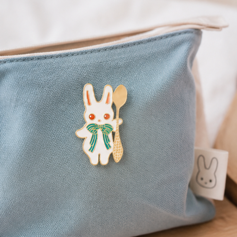 un pin's en forme de lapin blanc aux yeux rouges et qui a un joli noeud papillon vert. Il tient une cuillère dorée.