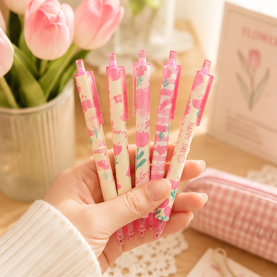 Main tenant un lot de stylos kawaii roses à motifs floraux pastel et tulipes, dans une ambiance douce et cosy avec fleurs roses, trousse assortie et décoration papeterie mignonne.