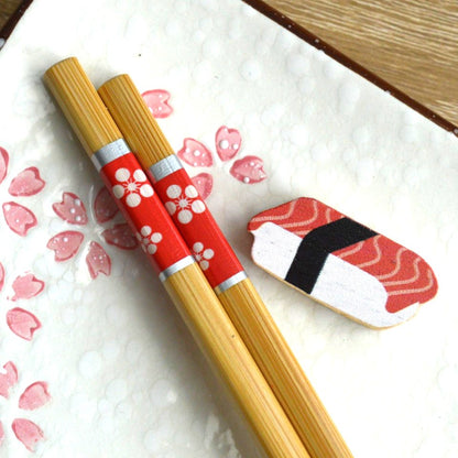 une paire de baguettes japonaises en bambou durable décorées de fleurs blanches sur un fond rouge. Elles ont un support pour baguettes en forme de sushi au thon