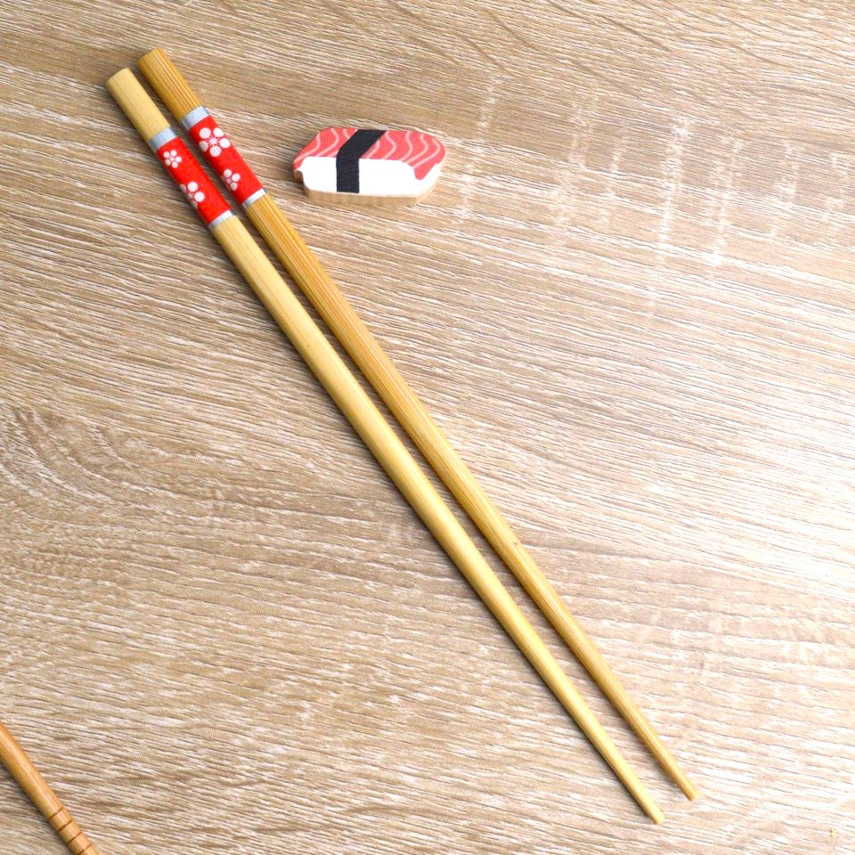 une paire de baguettes japonaises en bambou durable décorées de fleurs blanches sur un fond rouge. Elles ont un support pour baguettes en forme de sushi au thon