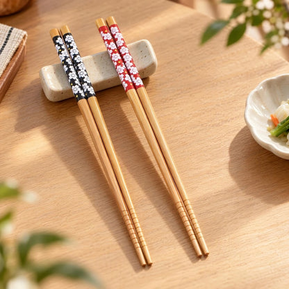 Deux paires de baguettes en bambou, une noire et une rouge, au motif de fleurs de sakura du Japon, posées sur un repose baguette sur une jolie table. Ambiance zen, calme qui rappelle le Japon.