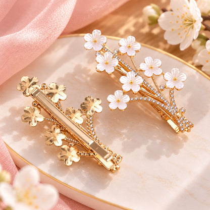 Deux vues d’une barrette à cheveux dorée en forme de branche de prunier, ornée de fleurs blanches nacrées et de strass brillants.