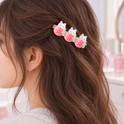 Barrette à cheveux kawaii ornée de trois adorables lapins blancs sur des fraises roses à pois blancs et feuilles vertes, portée sur le côté de la tête d’une femme aux cheveux bruns naturellement ondulés. Accessoire cheveux original et mignon, au style japonais kawaii, idéal pour une coiffure douce, fraîche et féminine.