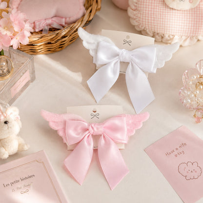 Deux barrettes à cheveux en forme de nœud avec ailes, l'une blanche et l'autre rose, présentées dans un décor kawaii aux tons pastel. Ces accessoires élégants et romantiques apportent une touche poétique et féminine à toutes les coiffures.
