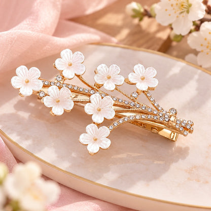 Barrette à cheveux dorée en forme de branche de prunier, ornée de fleurs blanches nacrées et de strass brillants, dans un décor romantique et élégant.