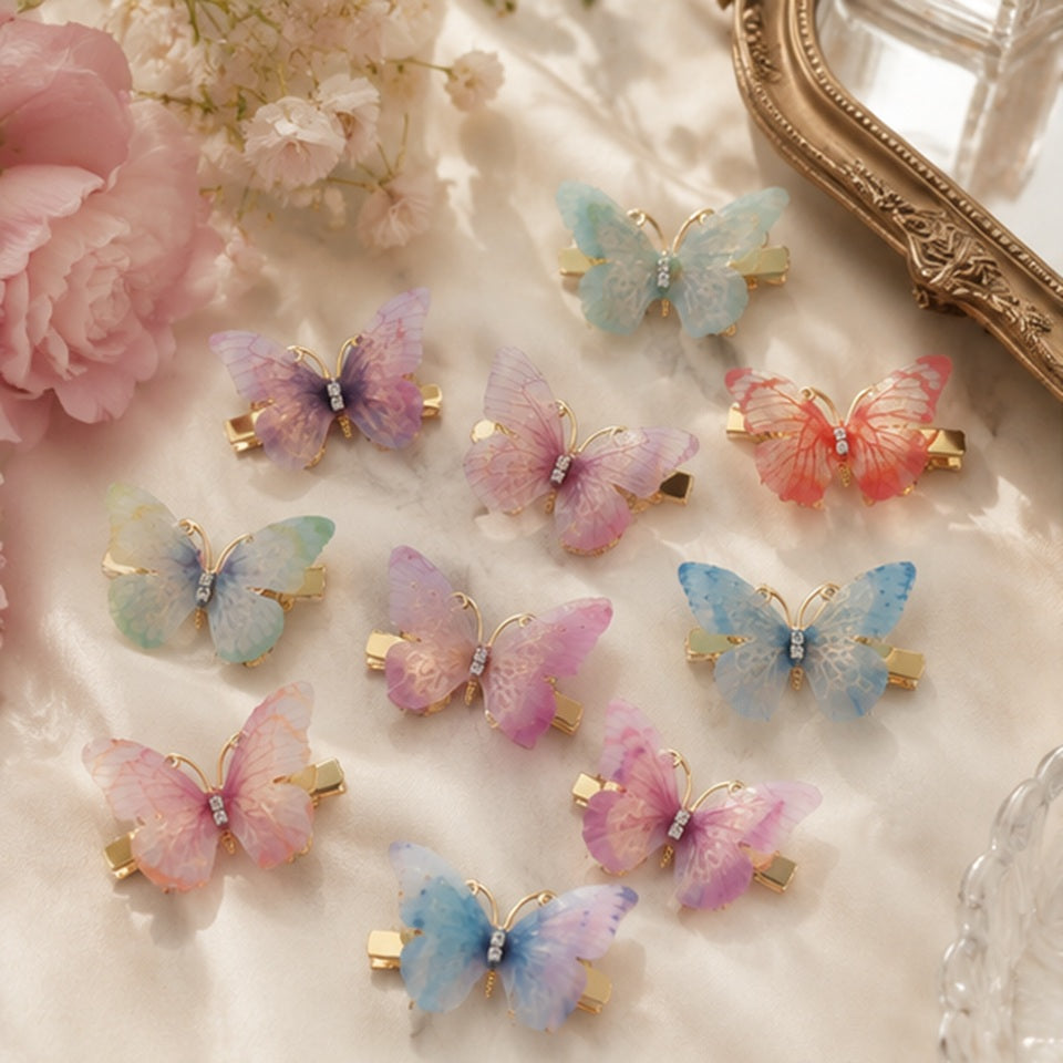 des barrettes à cheveux en forme de papillons colorés posés sur une coiffeuse avec des fleurs roses et un miroir. Les ailes des papillons sont en tissu colorés et ils sont dorés.