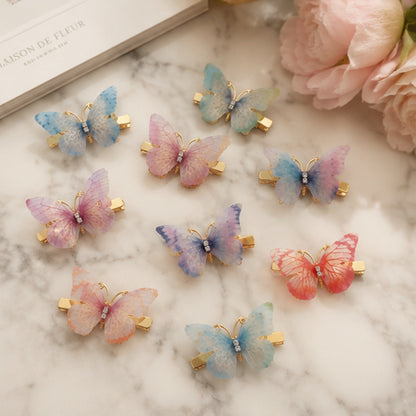 des barrettes à cheveux en forme de papillons colorés posés sur une table en marbre. Les ailes des papillons sont en tissu colorés et ils sont dorés.