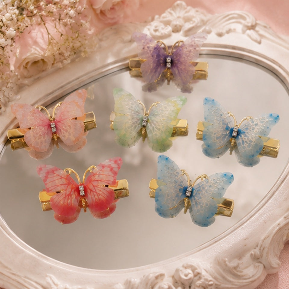 des barrettes à cheveux en forme de papillons colorés posés sur un petit miroir dans une ambiance feutré, romantique et printanière. Les ailes des papillons sont en tissu colorés et ils sont dorés.