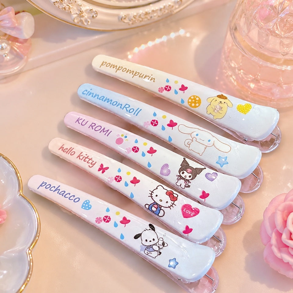 des barrettes à cheveux illustrées des personnages Sanrio : kuromi, hello kitty, pochacco, cinnamoroll et pompompurin dans des tons de couleurs pastels. Elles sont posées sur une coiffeuse rose et féminine.