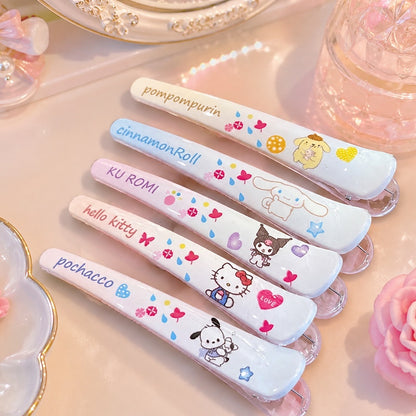 des barrettes à cheveux illustrées des personnages Sanrio : kuromi, hello kitty, pochacco, cinnamoroll et pompompurin dans des tons de couleurs pastels. Elles sont posées sur une coiffeuse rose et féminine.