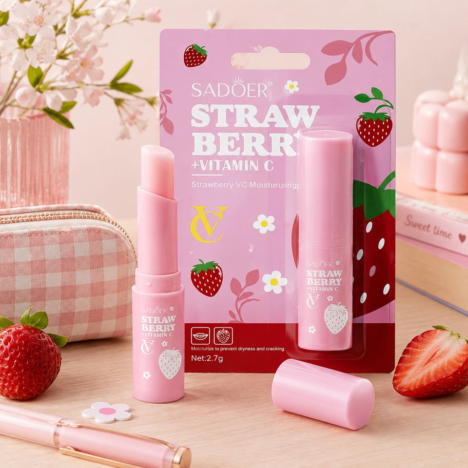 Baume à lèvres hydratant à la fraise et vitamine C SADOER, présenté avec son packaging rose, des fraises fraîches et des accessoires beauté dans un décor pastel.