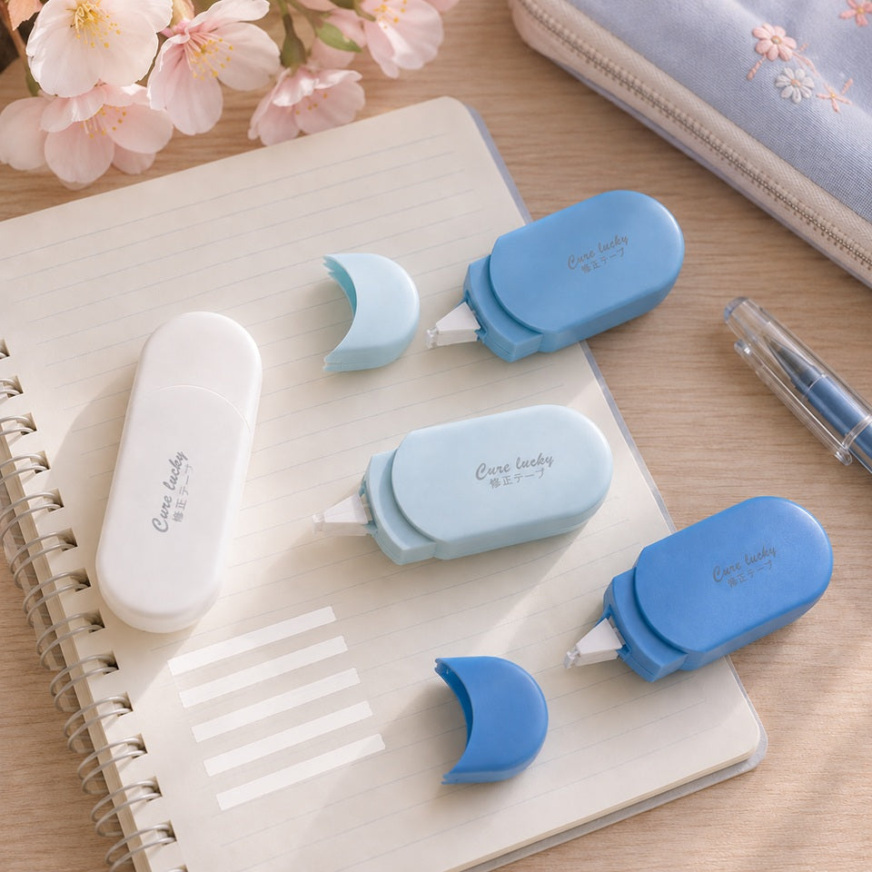 Blancos ruban correcteurs ovales en dégradé de bleu avec capuchons amovibles, présentés sur un carnet dans un joli décor de bureau avec papeterie et fleurs de sakura.