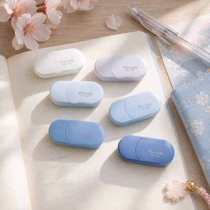 Blancos ruban correcteurs ovales en dégradé de bleu, présentés sur un joli carnet dans un décor de bureau lumineux avec papeterie et fleurs de sakura.