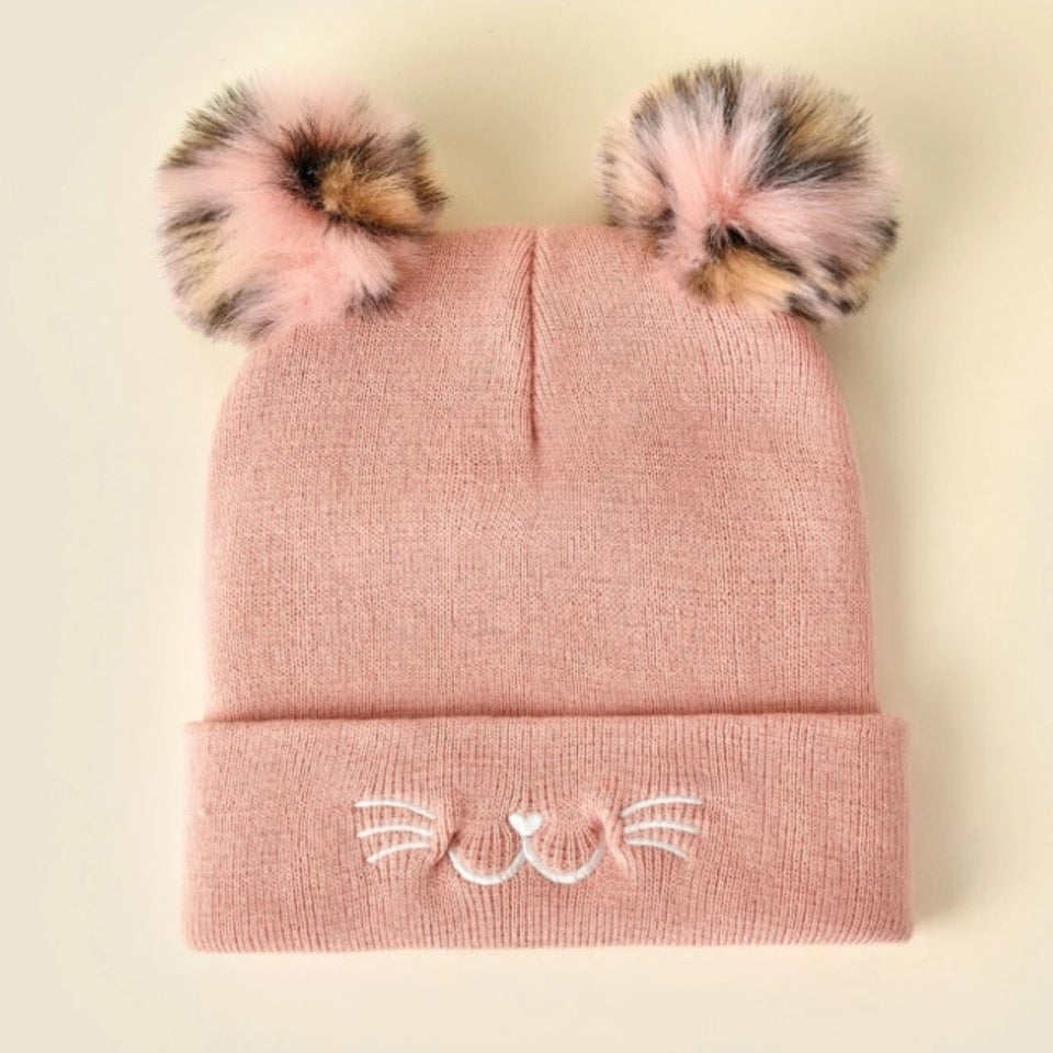 un bonnet rose tout doux avec deux pompons colorés. Sur le revers sont brodés des moustaches, une bouche et une truffe de chat. 