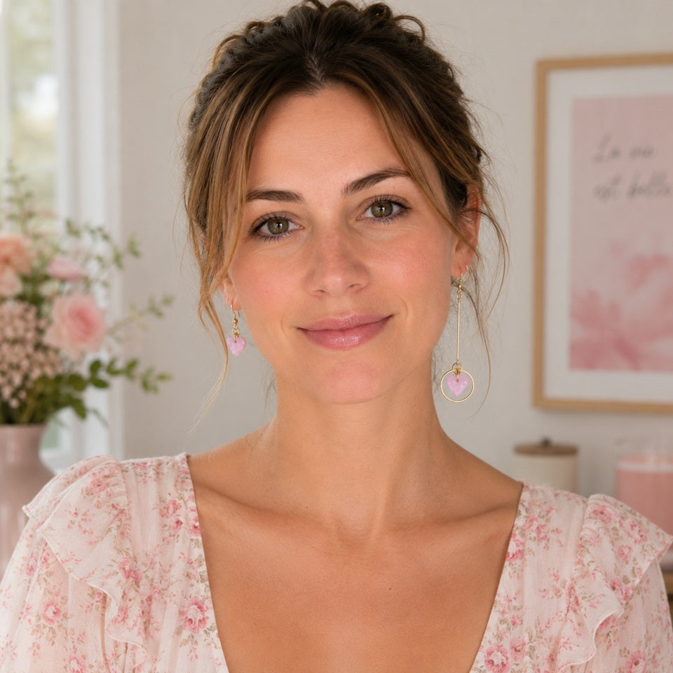 Femme portant des boucles d'oreilles asymétriques dorées avec pendentifs cœur rose nacré, vêtue d'une robe rose et blanche à fleurs, dans un décor pastel lumineux mettant en valeur ces bijoux fantaisie élégants et romantiques.