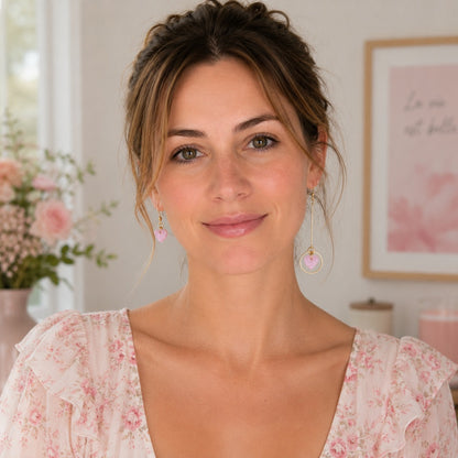 Femme portant des boucles d'oreilles asymétriques dorées avec pendentifs cœur rose nacré, vêtue d'une robe rose et blanche à fleurs, dans un décor pastel lumineux mettant en valeur ces bijoux fantaisie élégants et romantiques.