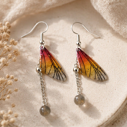 Boucles d'oreilles artisanales inspirées d'ailes de papillon aux dégradés orange, doré et gris, montées sur des crochets argentés. Chaque boucle est ornée d'une fine chaîne terminée par une perle grise translucide. Présentées sur une assiette en céramique beige avec un tissu naturel et des fleurs séchées, dans une lumière douce qui met en valeur les détails et les reflets.