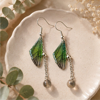 Boucles d'oreilles artisanales en forme d'ailes de papillon vert émeraude aux fines nervures noires, suspendues à des crochets argentés avec une chaîne délicate et une perle grise translucide. La mise en scène sur une assiette en céramique, entourée de feuilles d'eucalyptus, crée une ambiance naturelle, élégante et apaisante.