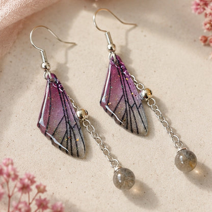 Boucles d'oreilles artisanales inspirées d'ailes de papillon aux nuances violettes et lilas légèrement pailletées. Les ailes sont associées à une chaîne argentée terminée par une perle grise translucide. Présentées sur une assiette en céramique beige avec un voile rose et de petites fleurs, elles sont sublimées par une lumière naturelle douce qui souligne leur finesse et leur éclat.