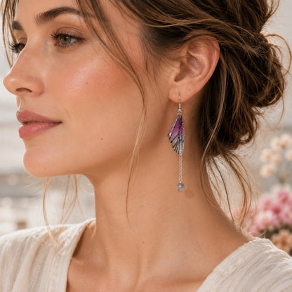 Gros plan d'une femme de type occidental portant une boucle d'oreille argentée composée d'une petite aile de papillon violette translucide aux reflets scintillants. Une fine chaînette pend verticalement derrière l'aile et se termine par une perle grise transparente. Les cheveux sont relevés en un chignon flou, le maquillage est naturel et l'arrière-plan, baigné d'une lumière douce, est délicatement flouté avec quelques fleurs pastel.