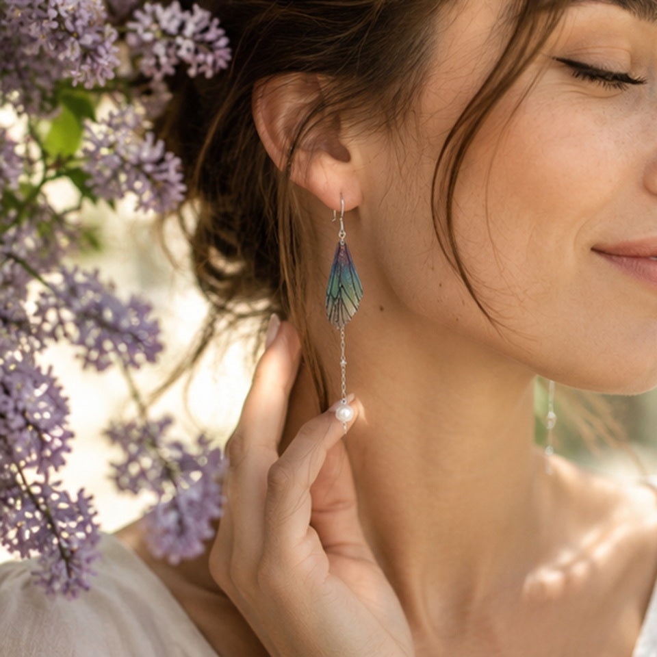 Gros plan sur une boucle d'oreille en forme d'aile de papillon irisée avec une fine chaîne et une perle blanche, portée par une femme dans un jardin fleuri aux tons lavande.
