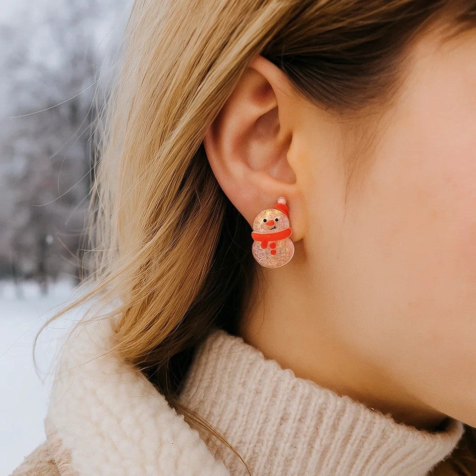 une paire de boucles d'oreilles en forme de bonhomme de neige qui porte un bonnet et une écharpe rouge. Ils sont tous pailletés et rappellent avec tendresse la brillance de l'hiver et de la neige.