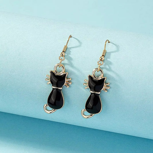 une paire de boucles d'oreilles dorées en forme de chats noirs, on voit leurs petites moustaches qui dépassent adorablement. Look mignon et tendre ! 