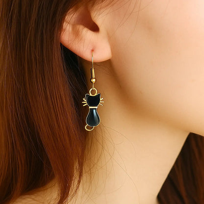 une paire de boucles d'oreilles dorées en forme de chats noirs, on voit leurs petites moustaches qui dépassent adorablement. Look mignon et tendre ! 