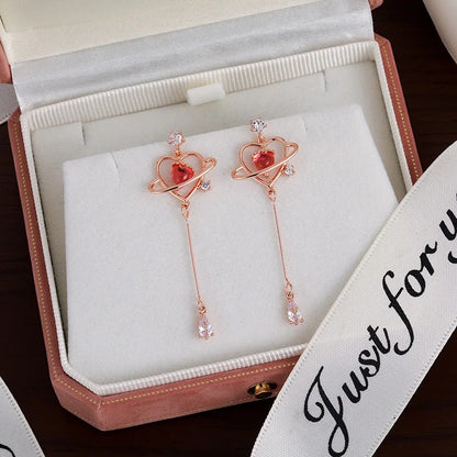 des boucles d'oreilles au design romantique et féminin : une structure en forme de coeur doré abrite une strass rose en forme de coeur, tel le coeur d'une planète. Au bout, est suspendu un strass brillant en forme de goutte.