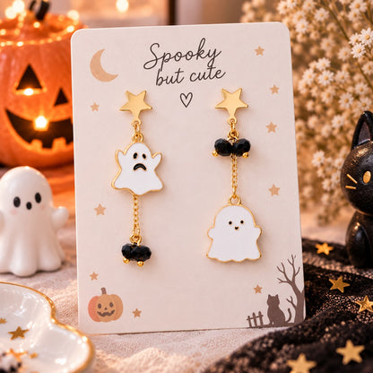 Boucles d’oreilles asymétriques fantômes kawaii dorées avec étoiles et perles noires, présentées dans un décor Halloween chaleureux avec citrouille et chat noir.