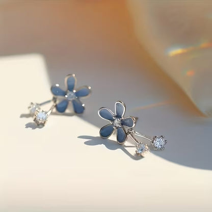 une paire de boucles d'oreilles composées d'une fleur bleue aux pétales arrondies d'où pendent deux strass brillants. l'ensemble est romantique et bohème