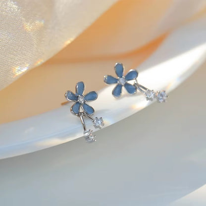 une paire de boucles d'oreilles composées d'une fleur bleue aux pétales arrondies d'où pendent deux strass brillants. l'ensemble est romantique et bohème