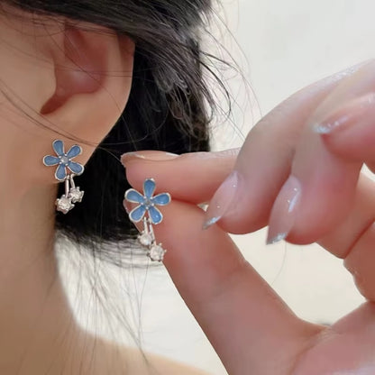 une paire de boucles d'oreilles composées d'une fleur bleue aux pétales arrondies d'où pendent deux strass brillants. l'ensemble est romantique et bohème