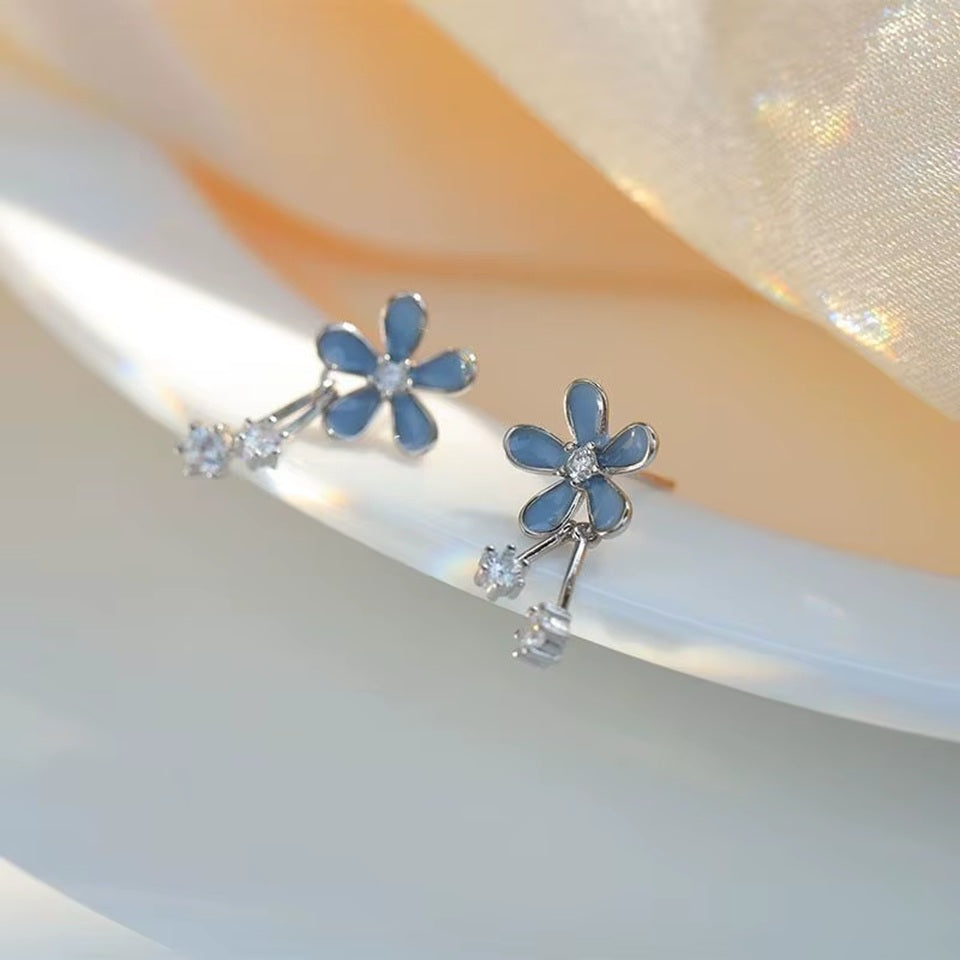 une paire de boucles d'oreilles composées d'une fleur bleue aux pétales arrondies d'où pendent deux strass brillants. l'ensemble est romantique et bohème