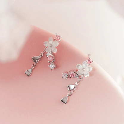 une paire de boucles d'oreilles qui épousent le lobe de l'oreille avec délicatesse et élégance. Une jolie fleur de sakura blanche et des strass blancs et roses viennent embellir l'ensemble. ne petite chaine pendante ornée de pétale de sakura est suspendue aux boucles.