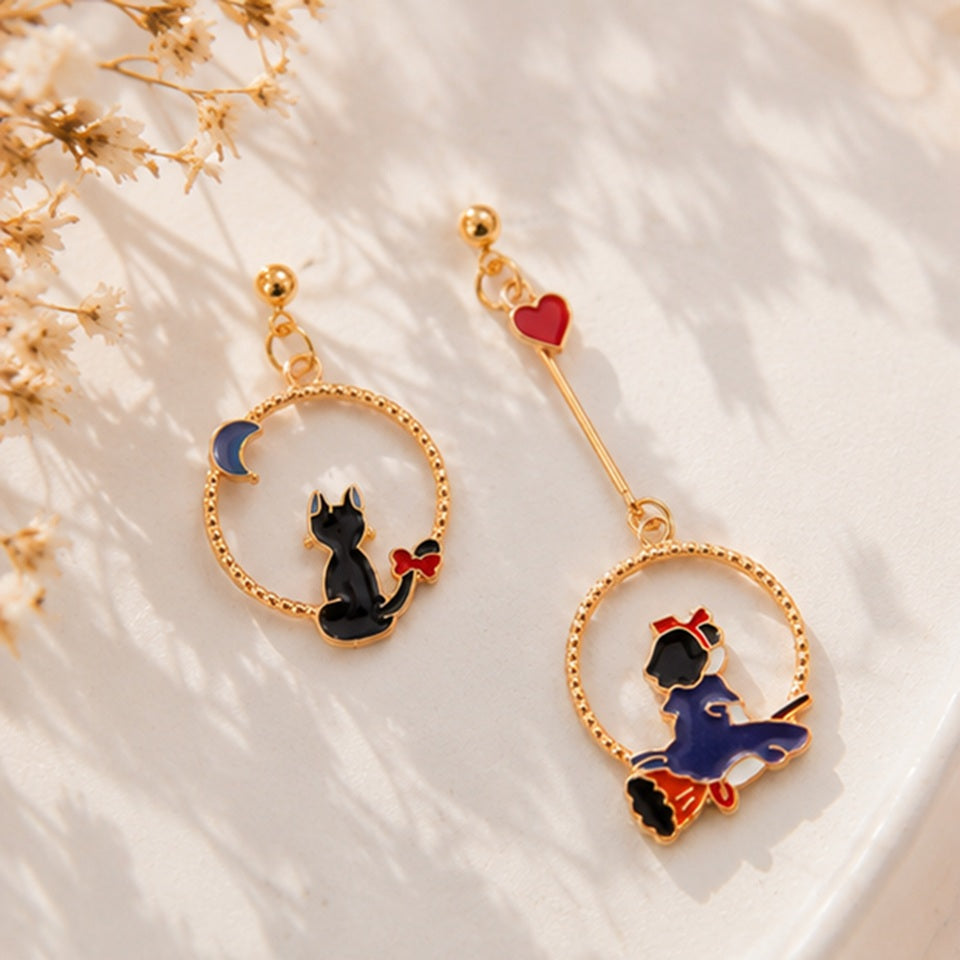 une paire de boucles d'oreilles asymétriques inspirées de Kiki la petite sorcière. D'un côté il y a Jiji le chat noir et de l'autre, Kiki sur son balai.